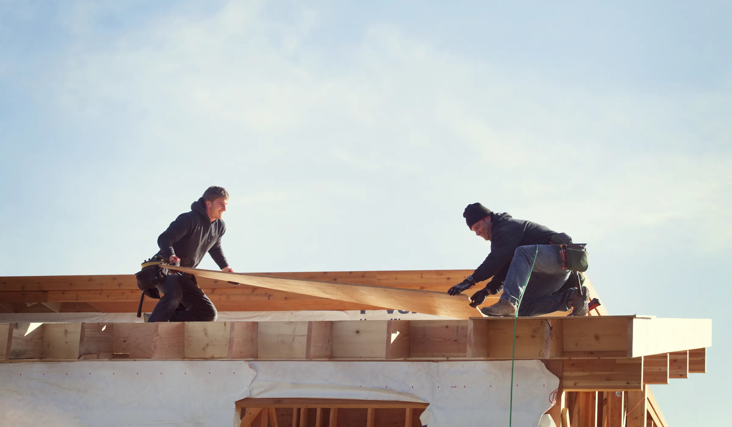 Deux charpentiers posent des pièces de bois sur la charpente d’une maison en construction, sous un ciel clair